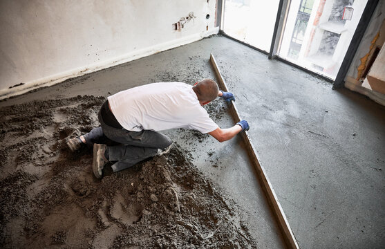 Male construction worker placing screed rail on the floor covered with sand-cement mix. Man smoothing and leveling surface with straight edge while screeding floor in apartment.