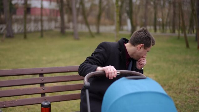 Young sick man with painful throat coughing sitting on bench in park shaking baby stroller. Portrait of unwell Caucasian father with newborn child outdoors