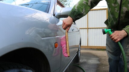 hands hold a brush with pink bristles and a green rubber hose with a spray nozzle and water...