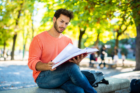 Handsome young man reading book on bench in the park - Powered by Adobe