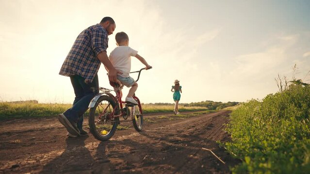 Mom dad teaching son ride bike in village on summer vacation. Parents running near child boy, mother father holding bike steering wheel supporting kid. Family weekend on nature, outdoors activity.
