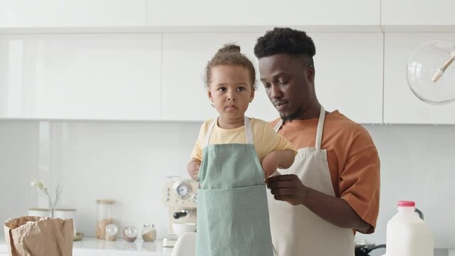 Young African American father helping little daughter to put on apron before cooking together in kitchen at home