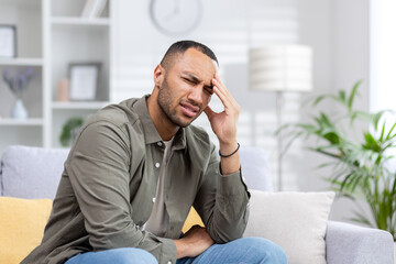 A young Muslim man is suffering from a headache. He sits on the sofa tiredly and massages his hand,...