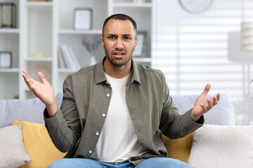 Dissatisfied and upset young African American man sitting at home on sofa in front of camera,...