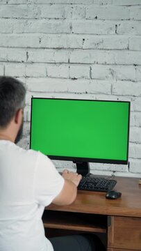 Bearded Middle-Aged Man Working at Desktop Computer With Green Mock Up Screen. Interior- Modern Office With White Brick Wall