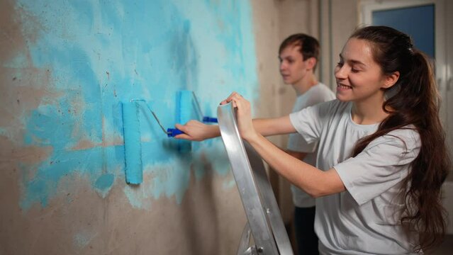 Happy couple in working clothes renovating new house apartment painting wall together in blue color using paint rollers. Home makeover, renovation concept. Family man woman repair flat themselves.