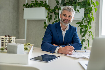 Man architect with beard working on some project, sitting at table in office in front of his laptop.