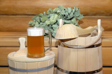 A glass mug of light cold beer stands on a wooden bucket near traditional bath accessories and dry oak whisk in a steam room of sauna.