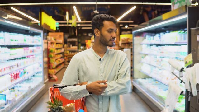 Smiling indian man walking in aisle with shopping basket at supermarket