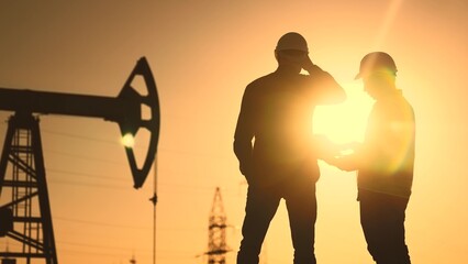oil production. two silhouette workers work as a team next to an oil pump. business oil production production concept. two engineers of the oil and gas industry are sun discussing a business plan - obrazy, fototapety, plakaty