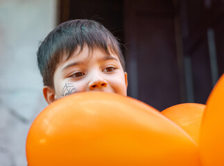 boy kid with orange balloons and spider web drawn on cheek playing outside, left building old door,...