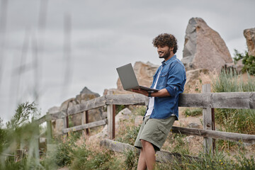 digital nomad concept, cheerful indian man standing near wooden fence and using laptop, nature