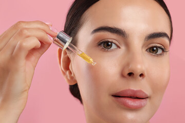 Obraz premium Beautiful young woman applying serum onto her face on pink background, closeup