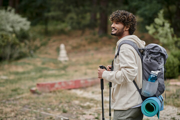 joyful indian backpacker holding trekking poles  and looking away in forest, adventure concept