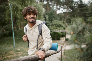 smiling indian tourist with backpack and smartwatch standing near fence in forest