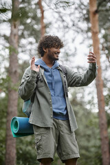 cheerful indian hiker with backpack having video call on smartphone in blurred forest