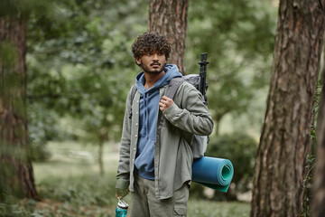 Fototapeta premium young indian traveler in shirt holding backpack and sports bottle near tees in forest