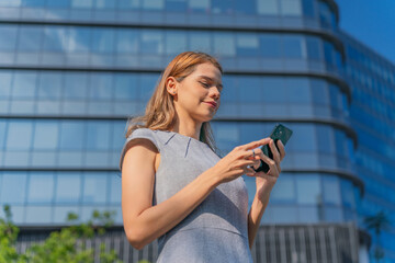 Business on the move. Young caucasian white businesswoman using smartphone while commuting in a central business district against a background of modern skyscraper cityscape.
