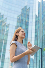Business on the move. Digital banking on the go. Portrait of a young and successful white caucasian business woman using digital tablet standing in front of a modern cityscape of office buildings. 