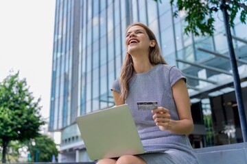 Young businesswoman doing digital banking transaction and making travel bookings using her credit card and her laptop in front a modern office building during her lunch break in a business district