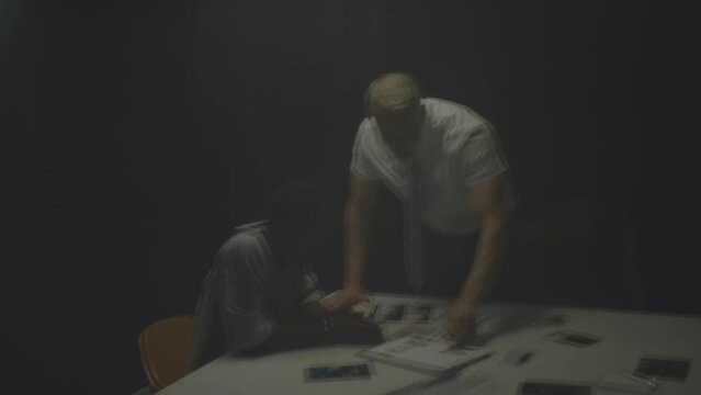 Through interrogation room window of mature FBI agent interviewing Black suspect man sitting in handcuffs at table
