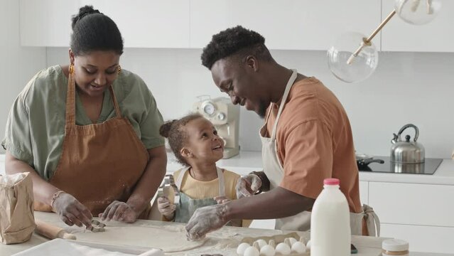 African American Parents And Their Little Daughter In Apron Cutting Cookies Out Of Dough While Baking Together In Kitchen At Home