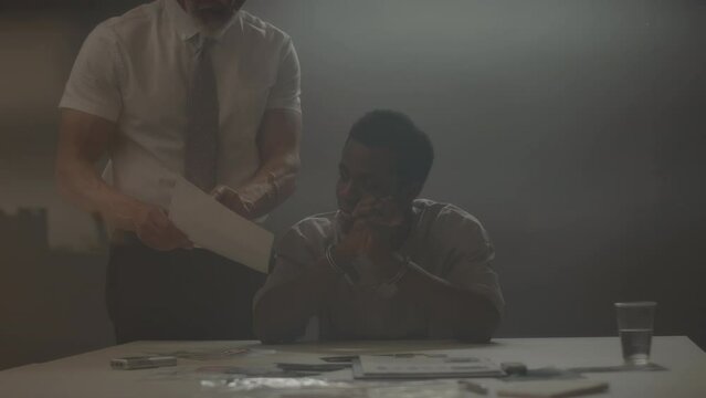 Waist up of suspect Black man being interviewed by police officer in interrogation room at night