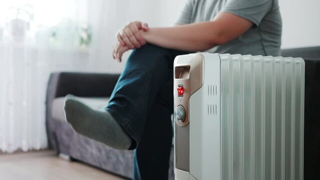 A man sits on the sofa in warm socks and turns on an electric heater to warm the apartment in cold weather. Copy space for text