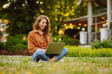 Young woman is sitting in a sunny park on a clearing and working with a laptop, blogging, online learning. Concept of remote work, freelancing, technology.