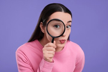 Confused young woman looking through magnifier glass on purple background