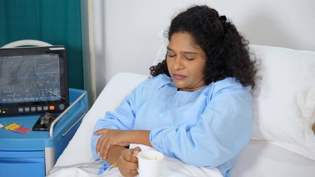 A middle-aged Indian woman sipping tea / coffee in a hospital room - hospital care  hospital food  ecg monitor. Asian female patient in blue hospital dress - hospital bed  sick lady  medical proble...