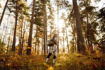 Tourist with a hiking backpack, hat walks along a path in the autumn forest. Beautiful woman enjoys...