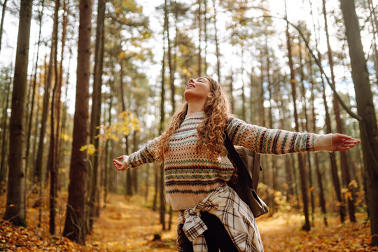 Tourist with a hiking backpack, hat walks along a path in the autumn forest. Beautiful woman enjoys a sunny day in nature, feels freedom and breathes fresh air, explores nature.