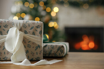 Stylish christmas gifts close up against tree lights bokeh and burning fireplace. Merry Christmas! Beautiful modern wrapped present with ribbon on wooden table. Christmas background, copy space