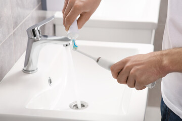 Man squeezing toothpaste from tube onto electric toothbrush above sink in bathroom, closeup
