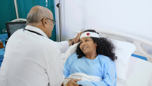 A senior male doctor consulting and consoling his female patient in the hospital ward - empathy  personal care . A middle-aged Indian woman with a head injury is recovering in the hospital - accide...