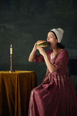 Junk food lover. Medieval woman in historical costume in image of renaissance maid eating burger...