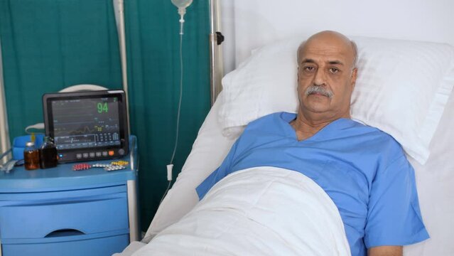An aged gentleman with a white moustache in the hospital bed - posing for camera  hospital care. Sick Indian old man wearing a blue hospital dress is admitted to the hospital - old age  medical pro...