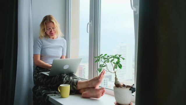 A happy barefoot blonde with a laptop, typing, smiling and drinking coffee, sitting on a windowsill in a cozy city apartment. Remote work, e-learning, the concept of freelancing.