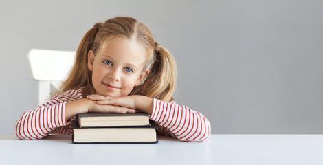 Banner of a beautiful little schoolgirl girl 6-7 years old looking at the camera, sitting and studying at table with books on a gray background. Copy space. Minimalism. Education and school concept.