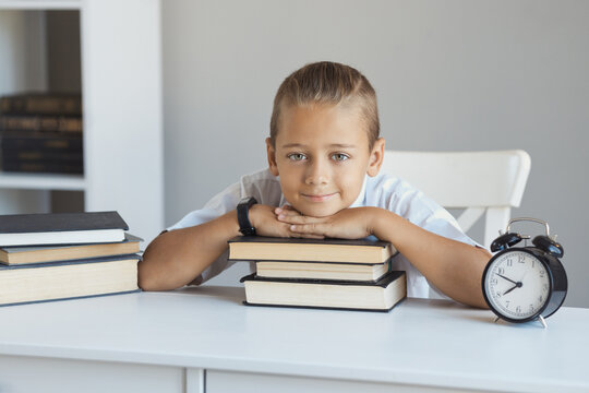Smart schoolboy in a white shirt sits at a desk with a large stack of books in the school library. Clever boy 8-9 years old doing homework and reading book at home.