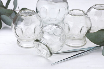 Glass cups, tweezers and eucalyptus leaves on white wooden table, closeup. Cupping therapy