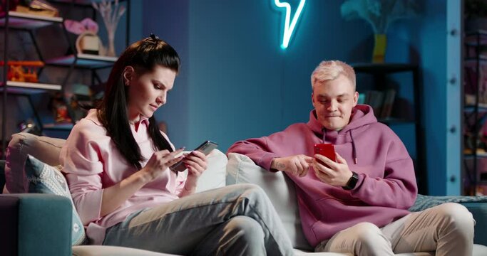 Fair-haired guy with dark-haired girl with phones on sofa in living room-studio. Unhappy and angry and happy, he won she lose. Relaxation recreation at free time. Modern leisure smartphone in internet