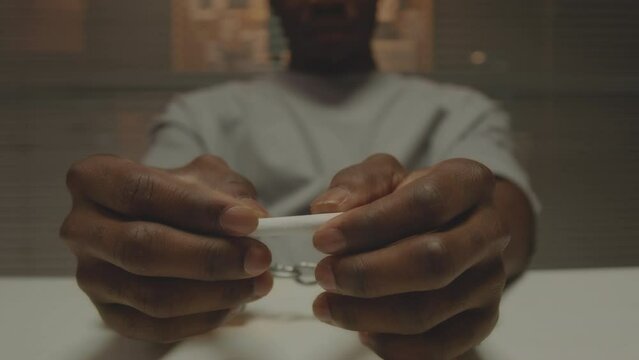 Closeup of African American criminal man in handcuffs holding cigarette in hands while sitting at table in dark interrogation room