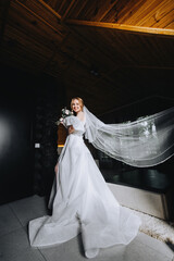 A beautiful young bride in a white long dress with a bouquet in her hands is standing in a room, indoors. Wedding photography, portrait, lifestyle.