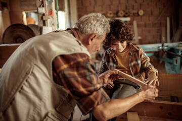 Grandfather carpenter teaching his grandson how to work with wood in a wood workshop