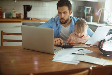 Young single father going over his bills and home finances while his daughter uses the tablet in the morning in the kitchen