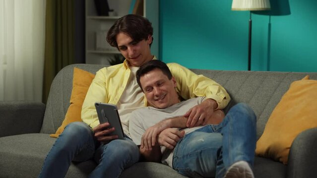 Video of a homosexual couple at home. They are laying on the couch, watching photos on a tablet, hugging, laughing and talking warmly to each other.