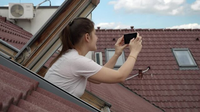 Young smiling woman looking out of the open attic window and making photos on her smartphone. Concept of tourism, holiday in Europe, travel and journey.