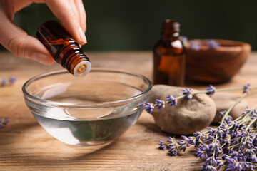 Woman dripping lavender essential oil from bottle into bowl at wooden table, closeup. Space for text